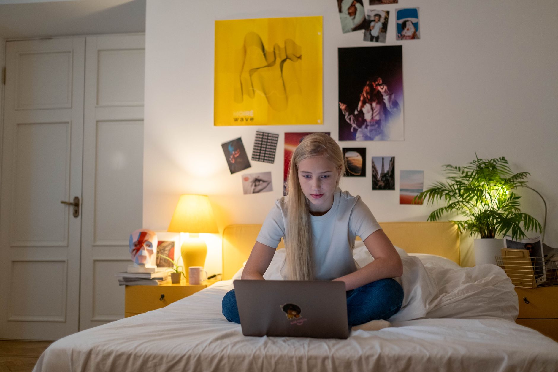 Una giovane seduta a gambe incrociate sul letto studia al portatile in una stanza illuminata da una lampada calda.